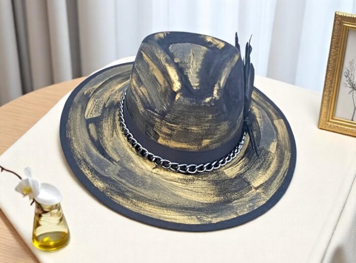 Power. Contrast. Mystery. This hand-painted hat blends deep black with bold golden strokes and a sleek metal chain for an edgy finish. One-of-a-kind, one-size (55–58 cm), crafted for those who turn every appearance into a statement of style.